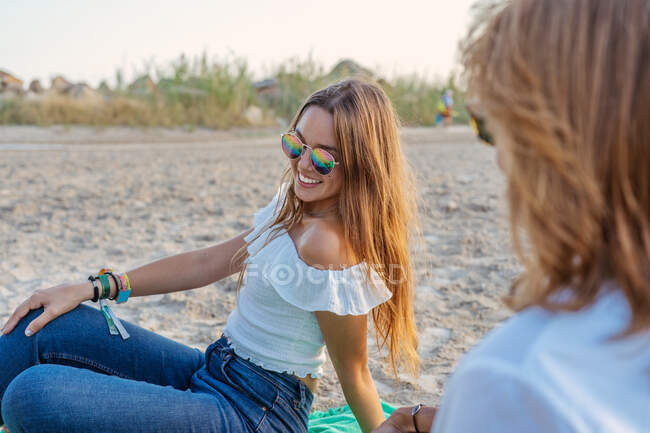 Vue latérale d'une jeune fille joyeuse souriante et communiquant avec une mère anonyme assise sur un rivage sablonneux lors d'une réunion de famille en station balnéaire — Photo de stock