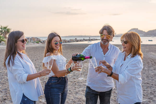 Alegre maduro padres y jóvenes hijas tintineo vasos de vino y riendo mientras se celebra la reunión familiar por la noche en el resort - foto de stock