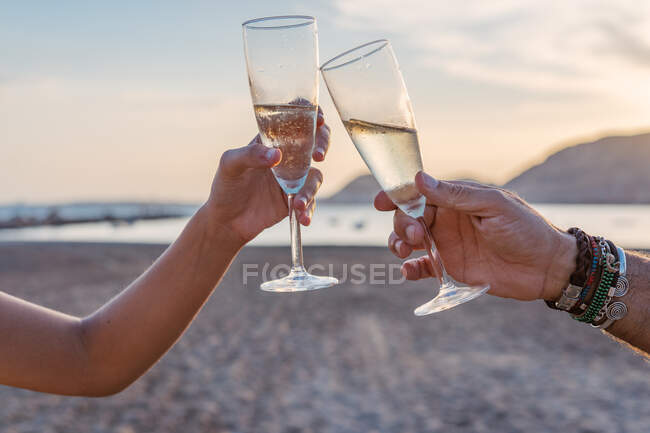 Madre e hija irreconocibles tintineando copas de vino y proponiendo brindis mientras celebran la reunión familiar en la playa de arena por la noche - foto de stock