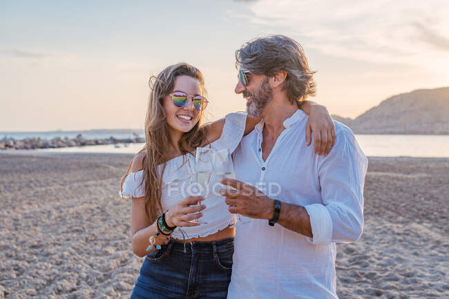 Homem maduro abraçando jovem mulher na bochecha enquanto propõe brinde e celebrando reunião familiar na praia de areia durante o crepúsculo no resort — Fotografia de Stock