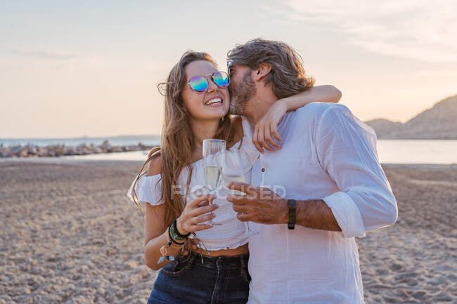 Reifer Mann umarmt und küsst junge Frau auf die Wange, während er ihr vorschlägt, anzustoßen und die Familienzusammenführung am Sandstrand in der Abenddämmerung auf dem Resort zu feiern — Stockfoto