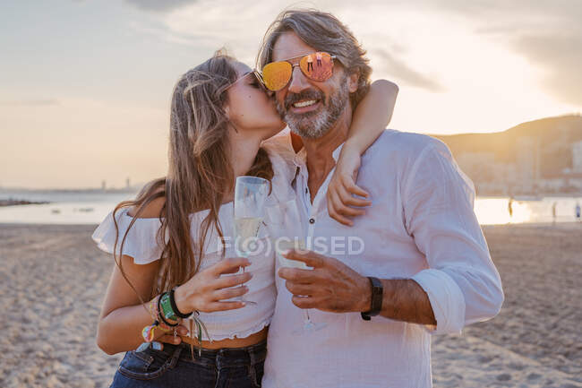Mature man hugging and kissing young woman in cheek while proposing toast and celebrating family reunion on sandy beach during dusk on resort — Stock Photo