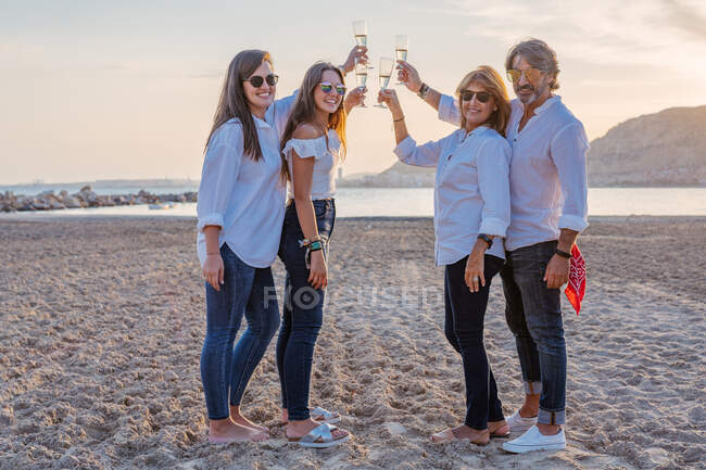 Cheerful mature parents and young daughters clinking glasses of wine and laughing while celebrating family reunion in evening on resort — Stock Photo