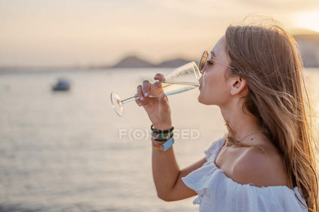 Side view of happy young female with closed eyes drinking wine while standing near sea in evening on resort — Stock Photo