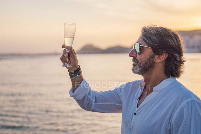 Side view of bearded mature man enjoying wine while standing against sea and sundown sky on resort — Stock Photo