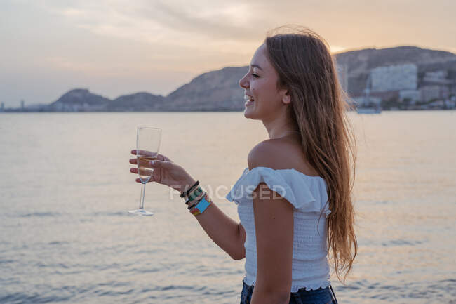 Jeune femme heureuse avec un verre de vin tout en se tenant près de la mer en soirée sur la station balnéaire — Photo de stock