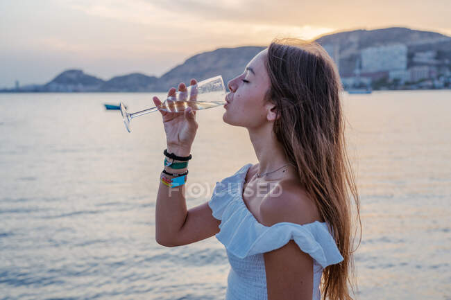 Seitenansicht einer glücklichen jungen Frau mit geschlossenen Augen, die Wein trinkt, während sie abends im Resort am Meer steht — Stockfoto