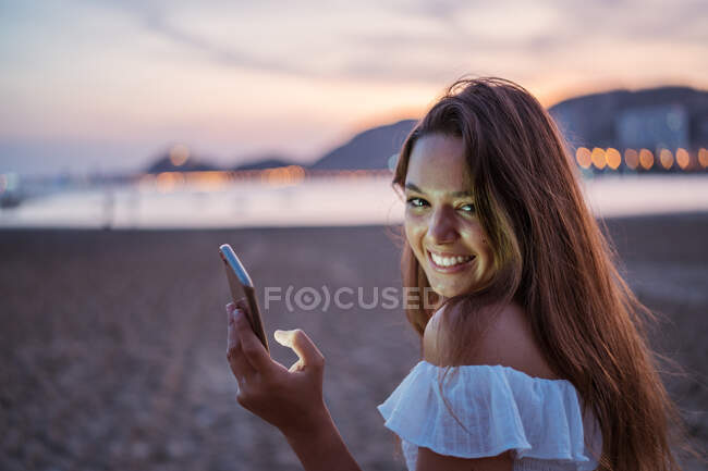 Visão traseira de uma jovem mulher feliz sorrindo e navegando nas mídias sociais no smartphone enquanto passa o tempo na praia de areia à noite — Fotografia de Stock