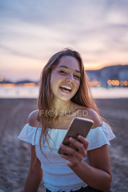 Feliz jovem mulher sorrindo e navegando mídias sociais no smartphone enquanto passa o tempo na praia de areia à noite — Fotografia de Stock