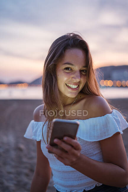 Happy young female smiling and browsing social media on smartphone while spending time on sandy beach in evening — Stock Photo