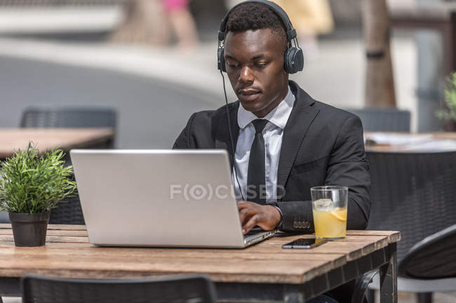 Homme noir en costume assis dans un café à l'extérieur avec ordinateur portable avec écouteurs filaires — Photo de stock
