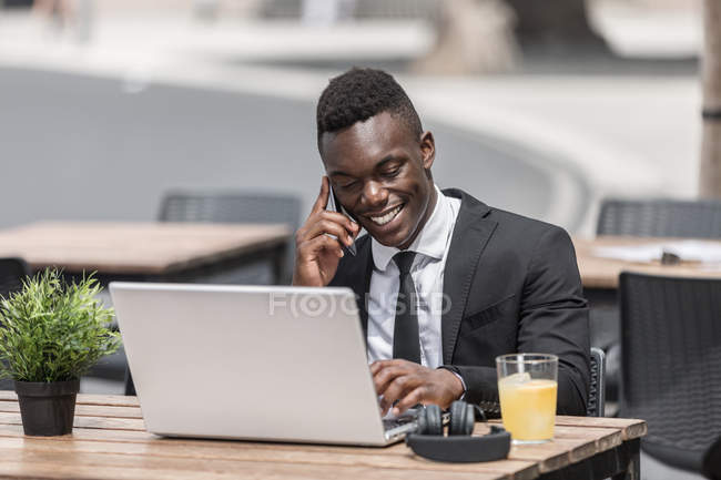 Afroamericano guapo freelancer hablando por teléfono mientras trabaja en el ordenador portátil en la cafetería urbana - foto de stock