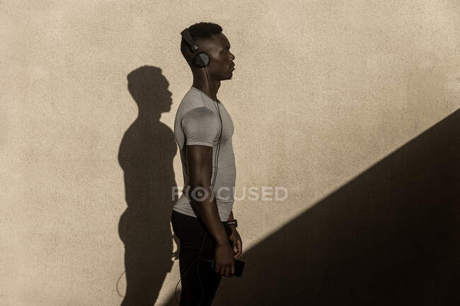 Vista lateral de atleta afroamericano adulto seguro en ropa deportiva y auriculares en el fondo de la pared gris con sombra durante el día - foto de stock