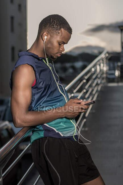 Deportista étnico complacido en auriculares que interactúan con el teléfono inteligente mientras se apoya en la cerca - foto de stock