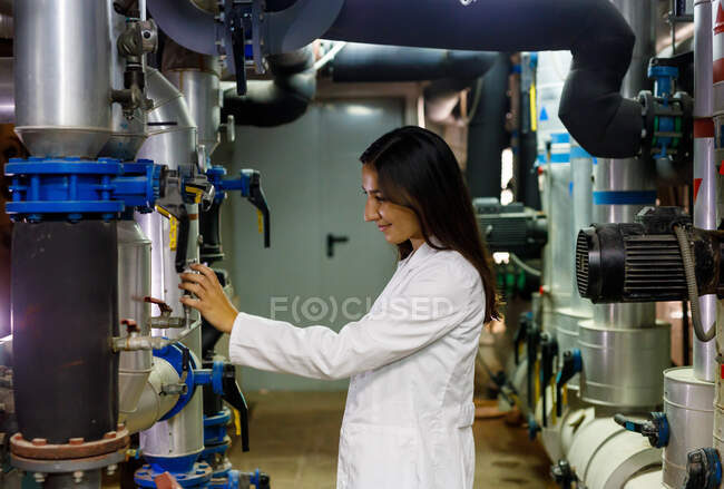 Vista lateral da jovem senhora positiva em válvulas de verificação de jaleco no sistema de tubulação em instalações industriais — Fotografia de Stock