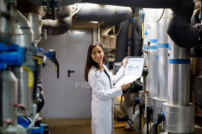 Side view of young woman in lab coat smiling and looking away while checking data in journal during safety inspection of piping system in industrial facility — Stock Photo