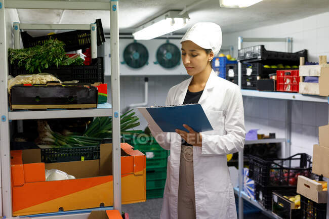 Woman in lab coat and hat making notes while checking food inside cold storage room in cafe — Stock Photo