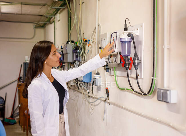 Vista lateral de una joven hembra con una bata de laboratorio presionando botones en un equipo industrial durante la inspección de seguridad en la instalación - foto de stock