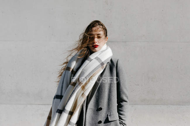Молодая женщина в стильном пальто и теплом шарфе стоит с закрытыми глазами у бетонной стены здания в ветреную погоду — стоковое фото
