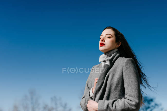 Vue latérale de la jeune femelle aux yeux fermés et au manteau élégant gris chaud debout contre un ciel bleu clair par temps venteux — Photo de stock