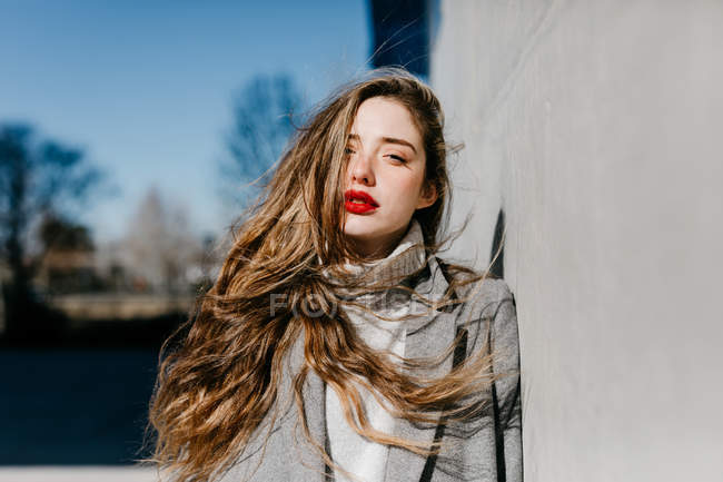 Привлекательная девушка смотрит в камеру, опираясь на бетонную стену здания в ветреную погоду — стоковое фото