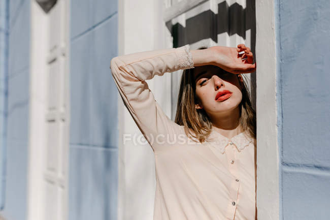 Young lady in elegant blouse covering closed eyes from sunlight while standing near white door of blue building on street — Stock Photo