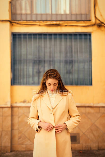 Sinnliche junge Frau mit langen Haaren, die stylische Jacke zuknöpft und nach unten schaut, während sie vor dem Gebäude an der Stadtstraße steht — Stockfoto