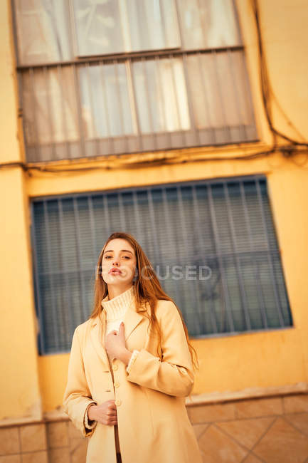 Чувственная молодая женщина с длинными волосами застегивает стильное пальто и смотрит в камеру, стоя снаружи здания на городской улице — стоковое фото