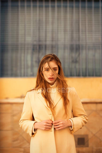 Sinnliche junge Frau mit langen Haaren, die stylischen Mantel zuknöpft und in die Kamera schaut, während sie vor dem Gebäude auf der Stadtstraße steht — Stockfoto