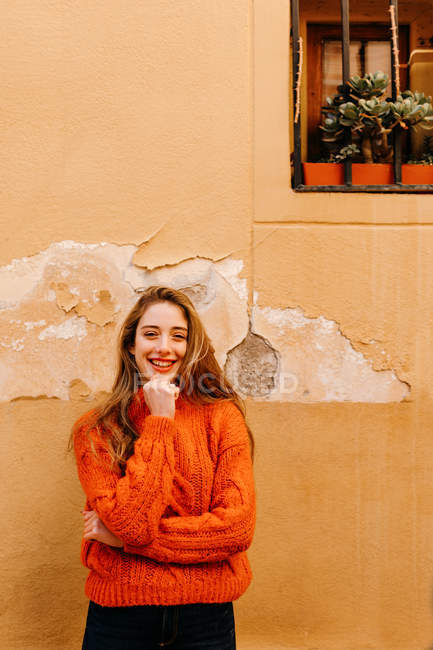 Чуттєва дівчина в модному в'язаному светрі торкається підборіддя і дивиться в камеру зовні будинку з горщиками на вікнах — стокове фото