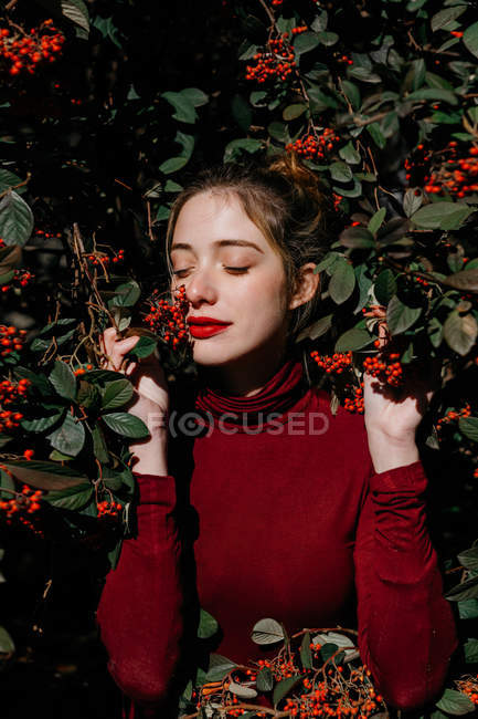 Jovem fêmea com olhos fechados em pé em meio a ramos verdes com bagas vermelhas no dia ensolarado no jardim — Fotografia de Stock