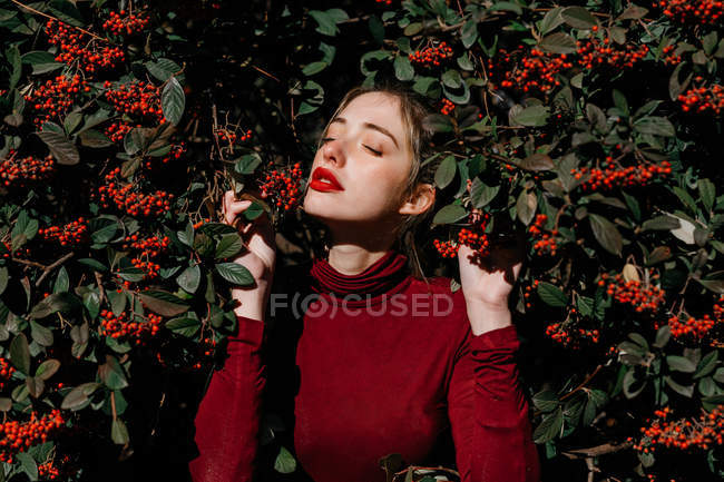 Молодая женщина с закрытыми глазами стоит среди зеленых ветвей с красными ягодами в солнечный день в саду — стоковое фото
