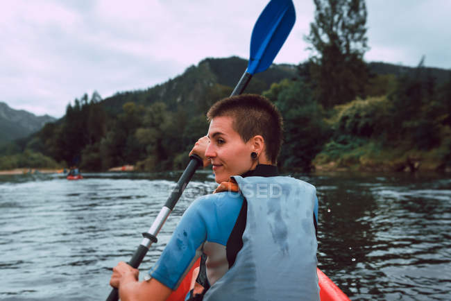 Vista posterior de la deportista mirando por encima del hombro mientras acolcha en canoa roja en el río Sella en España - foto de stock