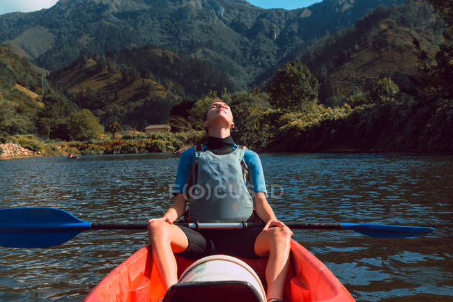 Mujer deportiva relajándose en canoa roja con los ojos cerrados en el declive del río Sella en España - foto de stock