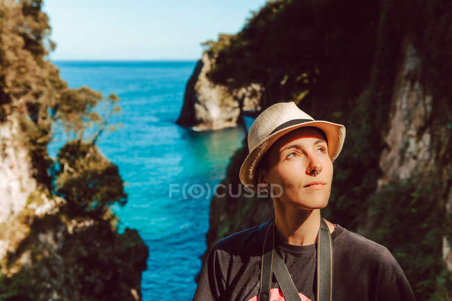 Young woman in hat standing with camera hanging on neck and enjoying picturesque view of sea and rocks in Ribadedeva Asturias Spain — Stock Photo