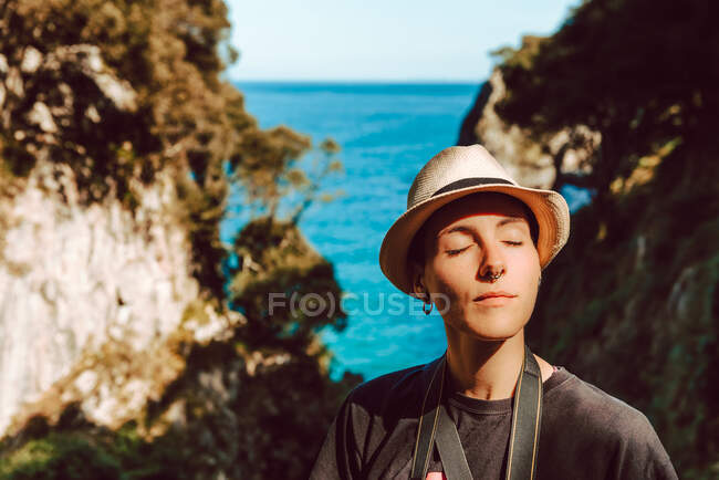 Mujer joven en sombrero de pie con cámara colgando del cuello y disfrutando de pintorescas vistas al mar y las rocas en Ribadedeva Asturias España - foto de stock