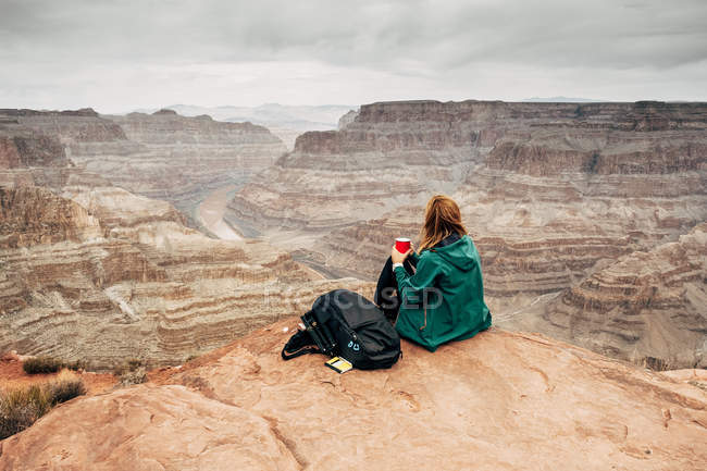 Vista lateral de mujer tranquila en chaqueta con mochila tomando café en las rocas y mirando el cañón en EE.UU. - foto de stock