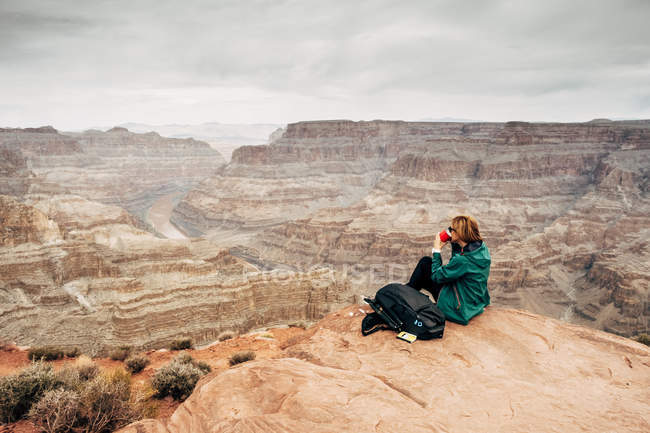 Vista laterale di donna tranquilla in giacca con zaino che prende il caffè sulle rocce e guardando canyon negli Stati Uniti — Foto stock