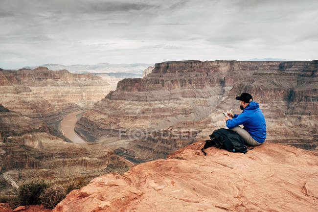 Vista lateral del hombre cansado relajándose en el borde de los acantilados en el cañón en EE.UU. y tomando notas en el cuaderno - foto de stock