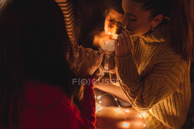 Deux amies en chandails buvant du chocolat chaud dans des tasses à Noël par la cheminée
. — Photo de stock