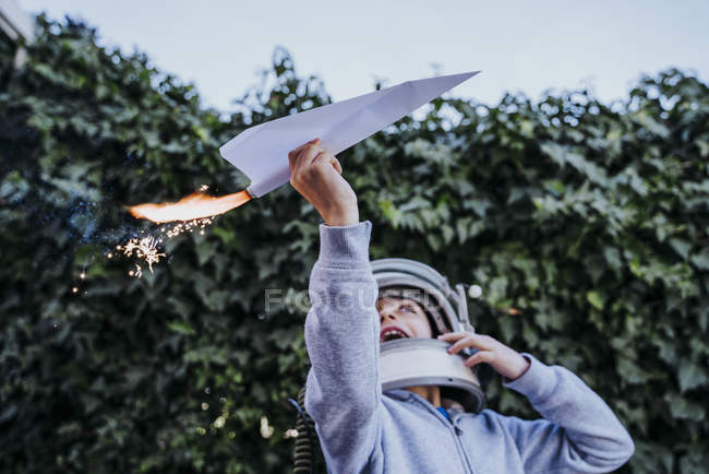 Возбужденный мальчик в шлеме астронавта играет с бумажным самолетом с петардой в саду — стоковое фото