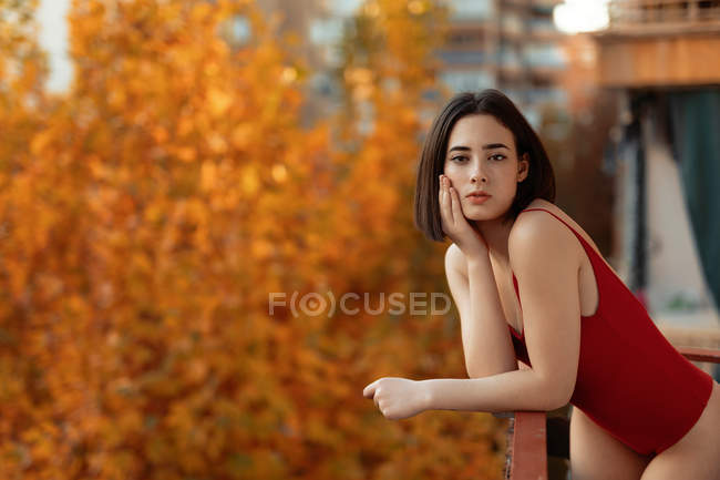 Linda jovem mulher em maiô vermelho inclinado sobre trilhos e olhando para a câmera com fundo embaçado outonal sazonal — Fotografia de Stock