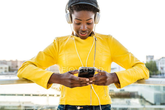 Femme afro-américaine dans une veste lumineuse élégante utilisant un téléphone portable et écoutant de la musique sur écouteurs — Photo de stock