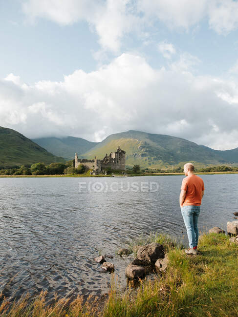 Side view of man standing by water enjoying picturesque landscape with green rocks and medieval Kilchurn castle on sunny day — Stock Photo