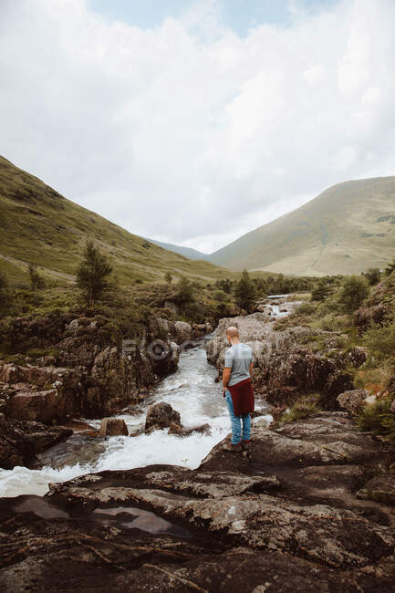 Seitenansicht eines männlichen Touristen, der auf Felsen über dem sprudelnden Fluss steht und tagsüber die Berglandschaft in Glencoe betrachtet — Stockfoto