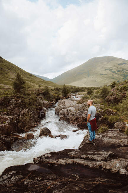 Vista lateral do turista masculino em pé sobre rochas acima do rio borbulhante e contemplando paisagem montanhosa em Glencoe durante o dia — Fotografia de Stock