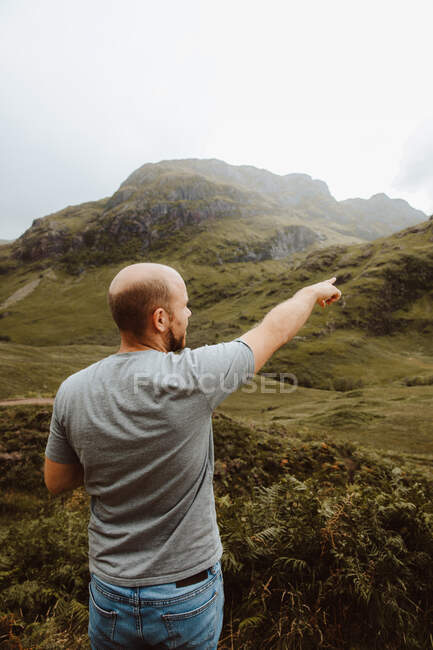 Vista posterior de calvo turista masculino de pie en el valle verde contemplando pintoresca vista y mostrando con el dedo índice en Glencoe - foto de stock