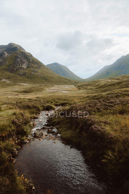 Ruhige Landschaft mit hohen Felsen, grünem Gras und kleinem Fluss unter bewölktem Himmel auf Glencoe — Stockfoto