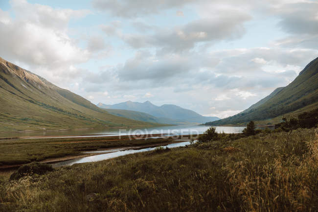 Paisaje idílico de altas montañas verdes y valle con un tranquilo río bajo cielo nublado en Glen en verano. - foto de stock