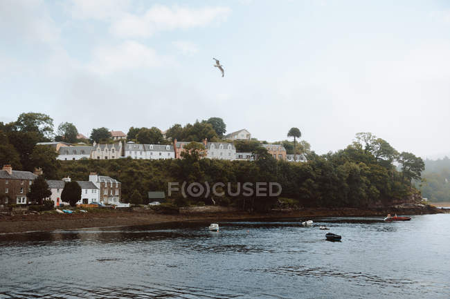 Düstere Landschaft verschiedener Häuser und Bäume auf einem Hügel am Wasser mit Segelbooten in Portree unter bewölktem Himmel — Stockfoto